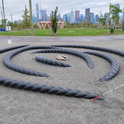 Image description: The image shows a walking labyrinth on gravel ground, marked by raised, twisted black textured guides forming concentric spiral paths. These guides help walkers follow the labyrinth's route with their feet or a cane. In the center of the labyrinth, there is a shell-shaped object. A white cane with a red tip is touching one of the black guides. Beyond the labyrinth, there is a grassy park with trees, a pedestrian path, and a skyline of tall city buildings under a cloudy sky. Photo by Leehe Lev.