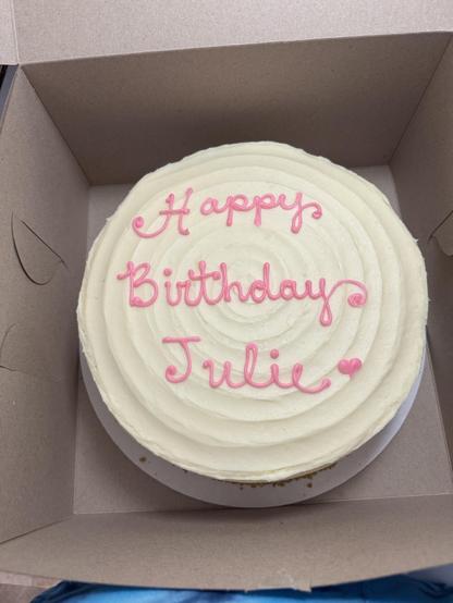 Round white-frosted cake in a cardboard box.  
The frosting is smooth with a spiral pattern from the center outward.  
Written on top in pink icing, in cursive, it says:  
“Happy  
Birthday  
Julie”  
There is a small pink heart shape after the name Julie.