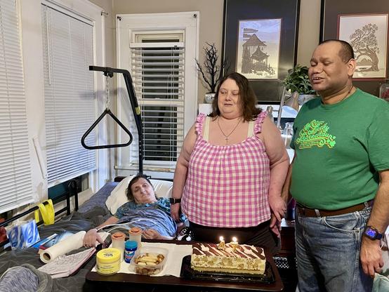 Three people in a room celebrating a birthday.  
In the center foreground is a rectangular birthday cake on a small table or tray, with white frosting, chocolate drizzle in a zigzag pattern, and two lit candles shaped like the numbers 4 and 7.  
Behind the cake stands a woman with light skin and shoulder-length brown hair, wearing a pink-and-white checkered sleeveless top; she has a necklace with a small cross and is facing slightly toward the right with a neutral, relaxed expression.  
To her right stands a man with medium-brown skin, short hair, and a green T‑shirt with yellow and white writing and shamrock graphics; he is wearing jeans and a smartwatch with a purple band, and his eyes are closed as though caught mid-song or mid-speech, with a slight smile.  
To the left, another woman with light skin and dark hair is lying in a bed; she is partially propped up on pillows, covered with a blue patterned blanket, and wearing a blue top. Her right arm rests on the bed’s side tray, near various small containers and a white paper towel roll.  
Above the bed is a black metal trapeze bar hanging from a frame.  
The room has light-colored walls and several windows with white blinds along the left side; behind the standing pair are framed artworks featuring landscapes and trees, and a decorative bare-branch ornament on a shelf.  
The overall scene feels like an intimate indoor birthday celebration near a bedside.